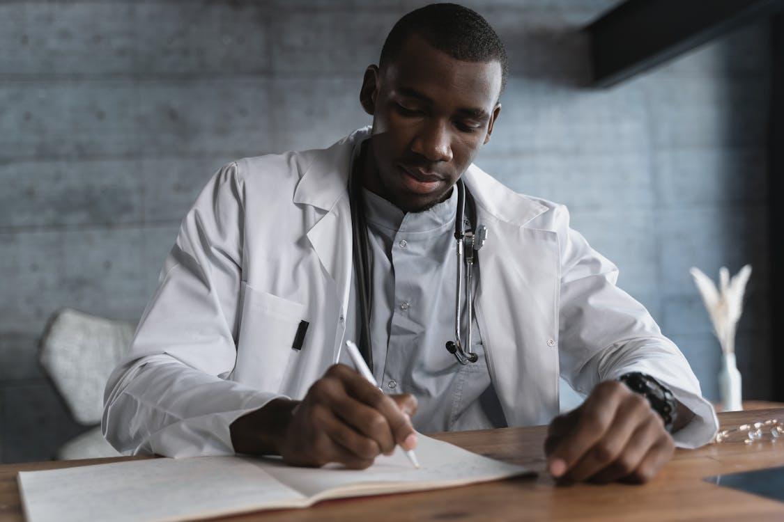 A doctor in a white coat sits at a desk, focused on writing a formal note or certificate on a document.