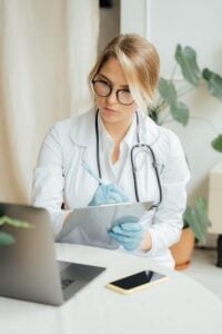 A doctor in a lab coat looks at a laptop screen while writing notes on a clipboard.
