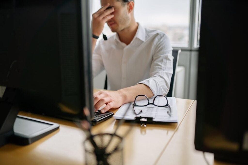 A man in a white shirt sits at a desk using a desktop computer, appearing distressed.
