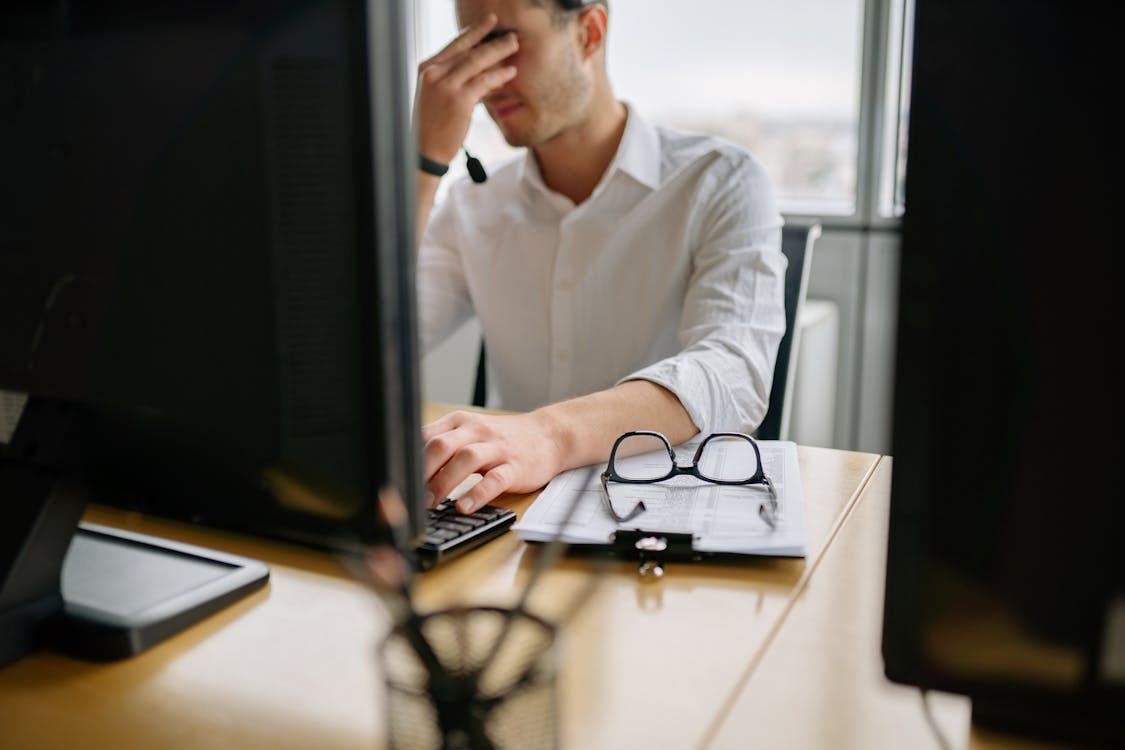 A man in a white shirt sits at a desk using a desktop computer, appearing distressed.