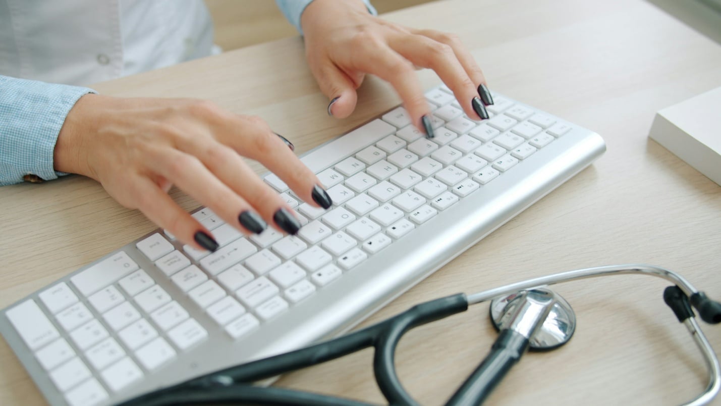 Hands typing on a keyboard next to a stethoscope while preparing medical documentation