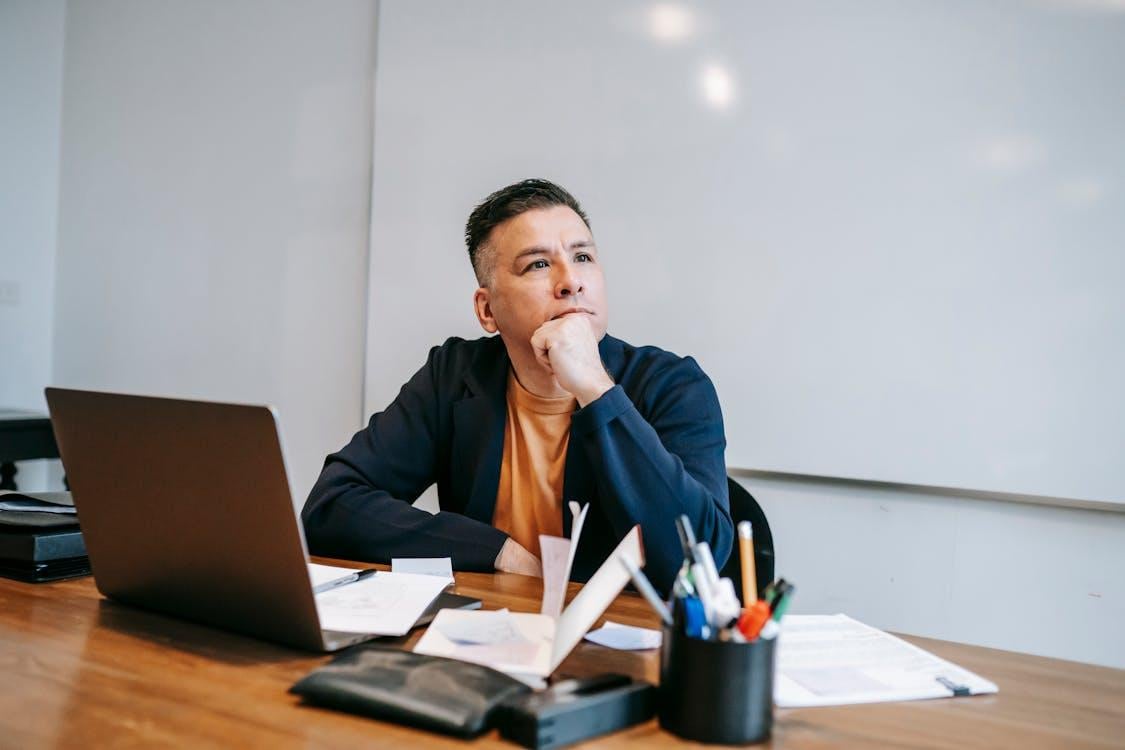 A man sits at a wooden desk, resting his chin on his hand.