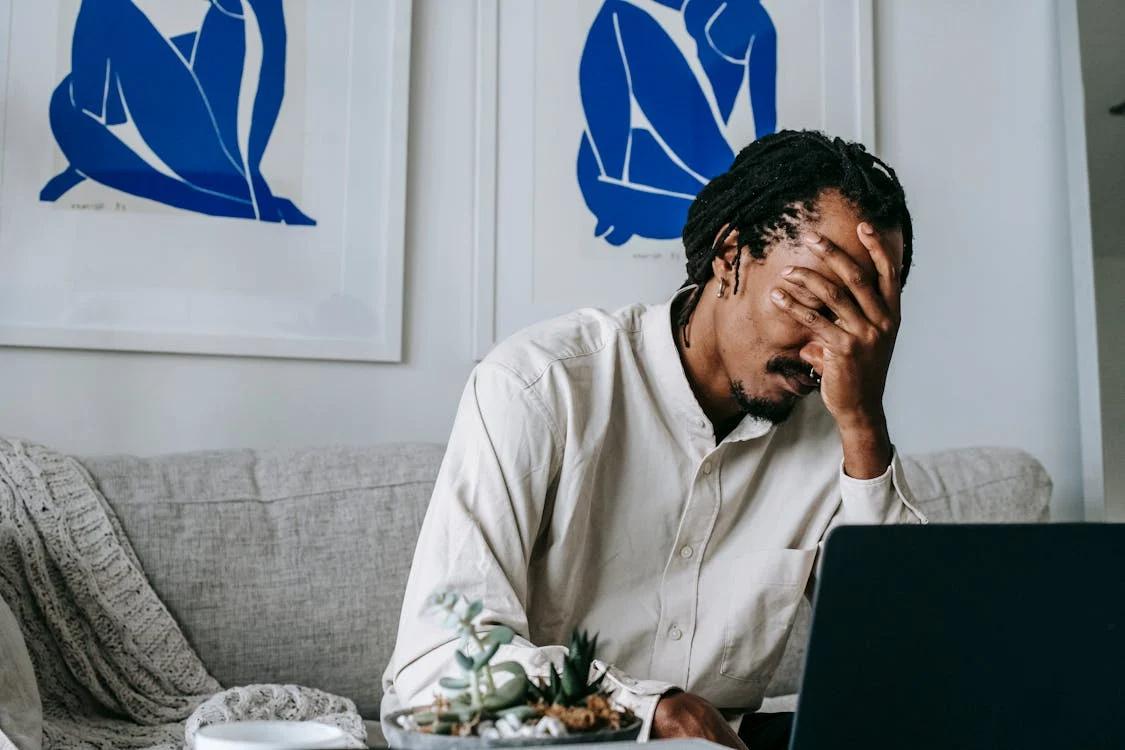 A young Black man looking visibly upset and covering part of his face with his hand.