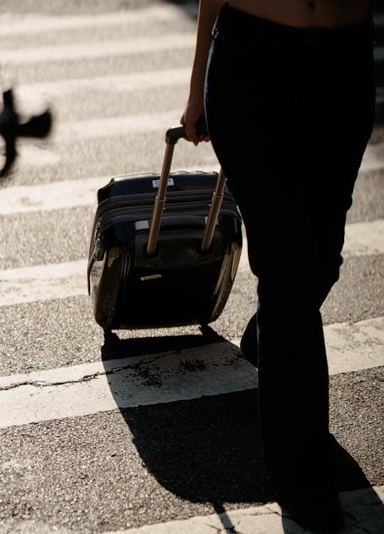 A close-up view of a person’s legs and a rolling suitcase as they walk across a marked street crossing in a city.