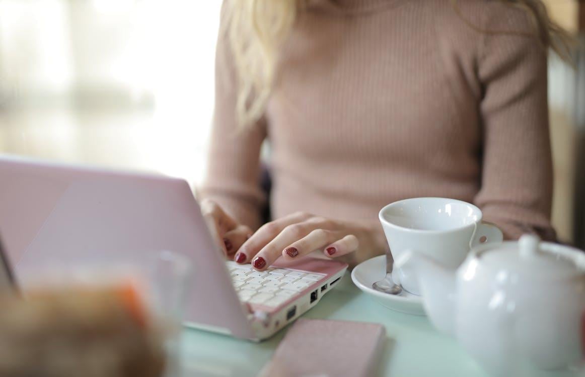 Close-up of hands typing on a laptop keyboard with a white ceramic mug of tea on a wooden desk.